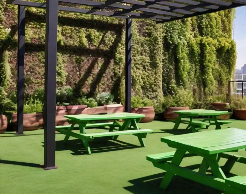 Green picnic tables and planters are arranged on a rooftop with a lush vertical garden and a pergola providing partial shade.